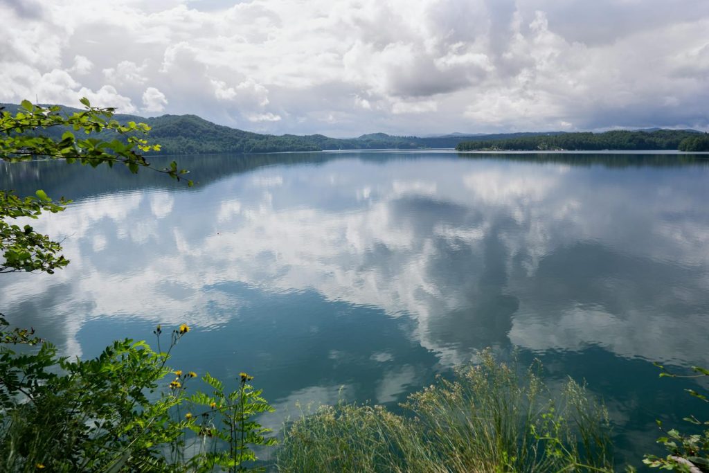 Serene lake landscape with clouds reflecting on calm water surrounded by lush greenery and distant hills