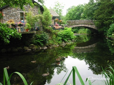 Idyllic stone cottage by a tranquil river with ducks and a charming arched bridge surrounded by lush greenery