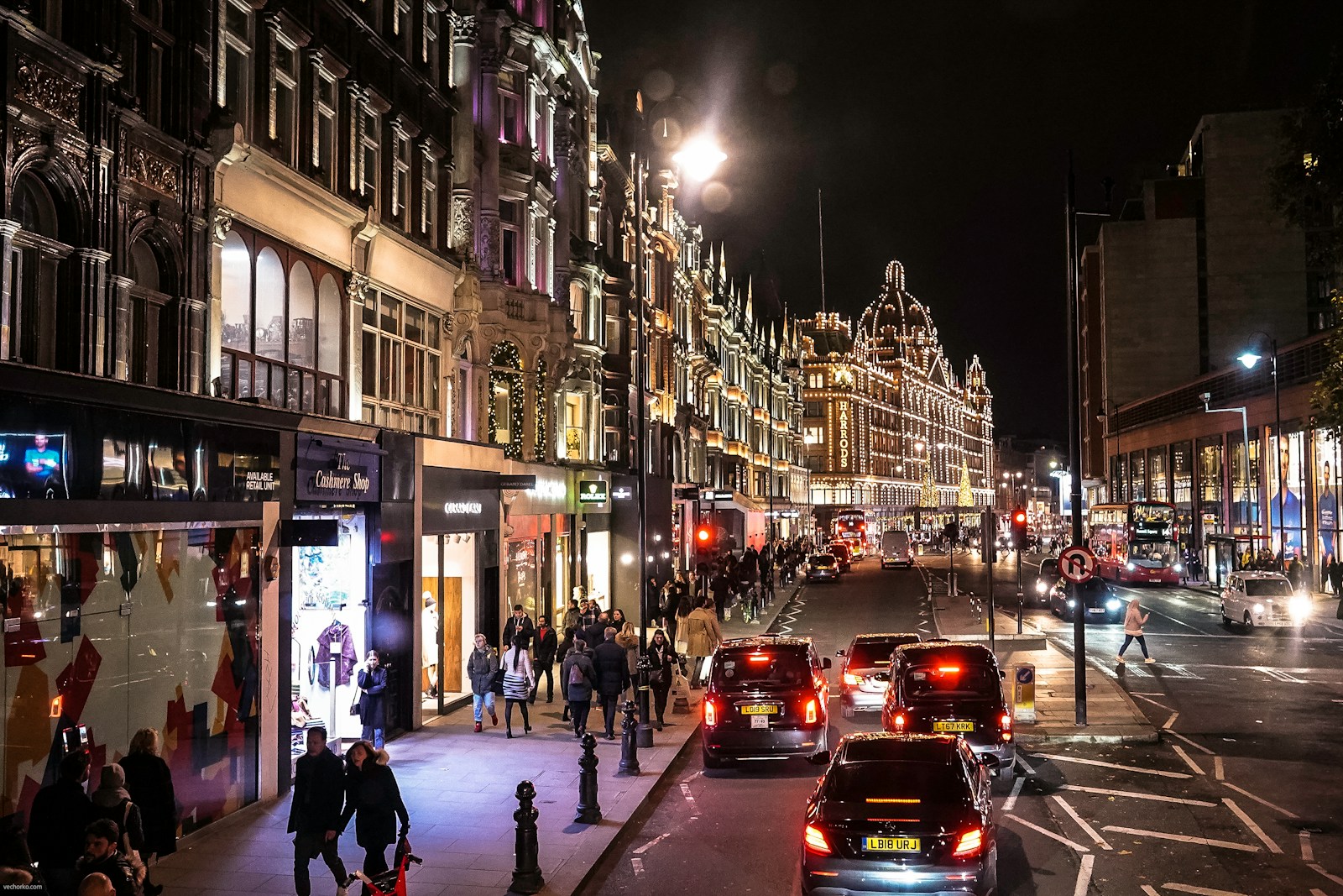 Busy London street at night with red buses and bright shopfronts, featuring iconic Harrods department store