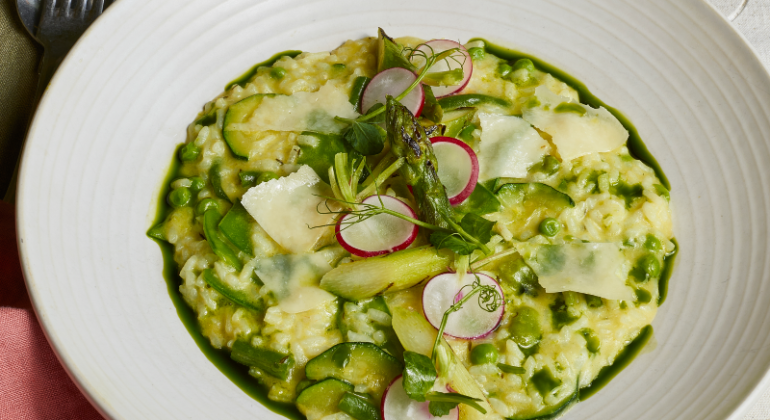 Creamy risotto with asparagus, peas and radishes served with a pink cocktail on a linen table setting