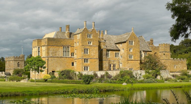 Historic English manor house by a pond under cloudy skies, surrounded by lush greenery and gardens