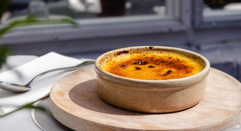 Crème brûlée in a ceramic dish on a wooden board with spoon and napkin in a bright setting