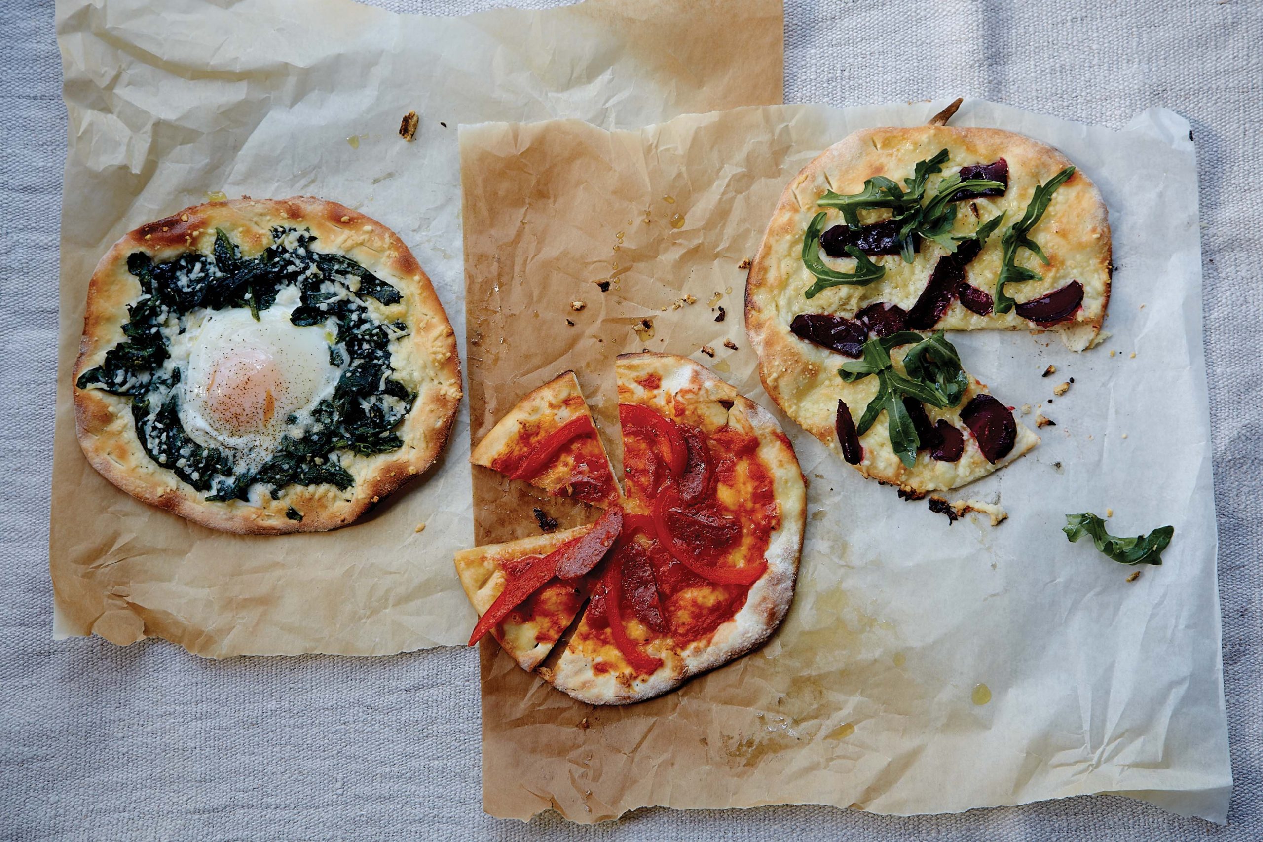 Three gourmet pizzas on parchment paper with diverse toppings including spinach, egg, peppers, and arugula.
