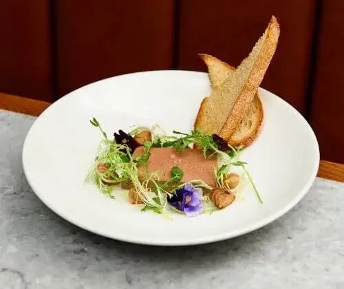 Gourmet pâté dish with crusty toast, garnished with edible flowers, greens, and nuts on a stylish white plate