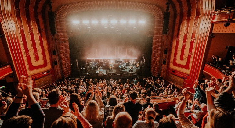 Crowd applauding at live concert in ornate theatre with bright stage lights