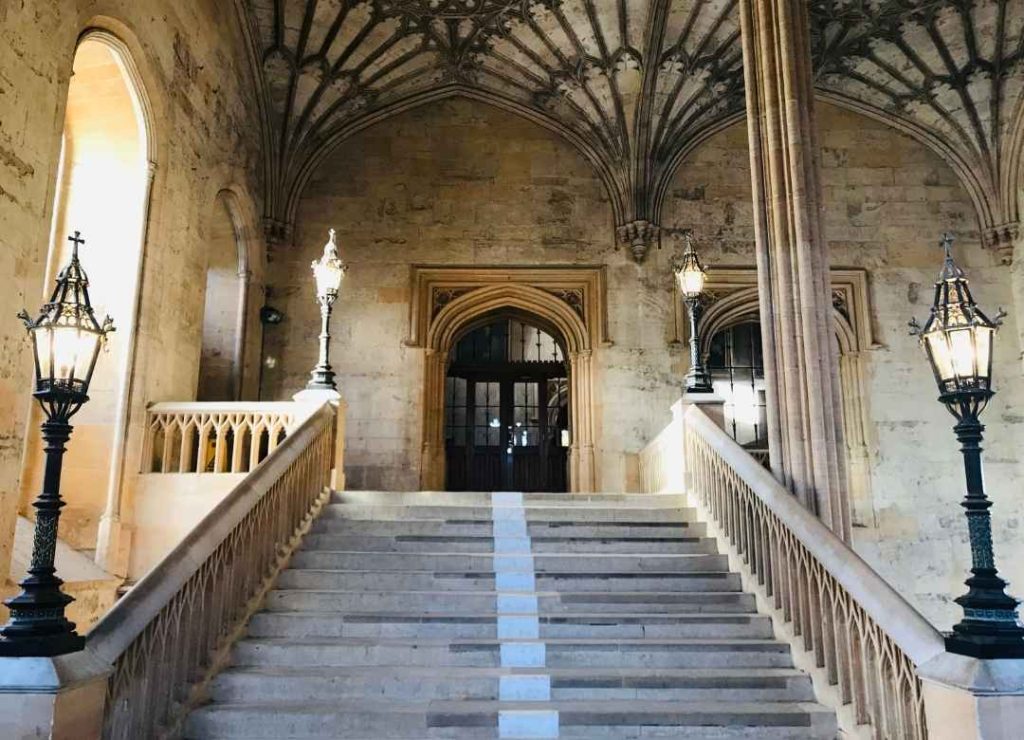 Ornate stone staircase with vintage lamps leading to arched wooden doors in historic building interior with vaulted ceiling