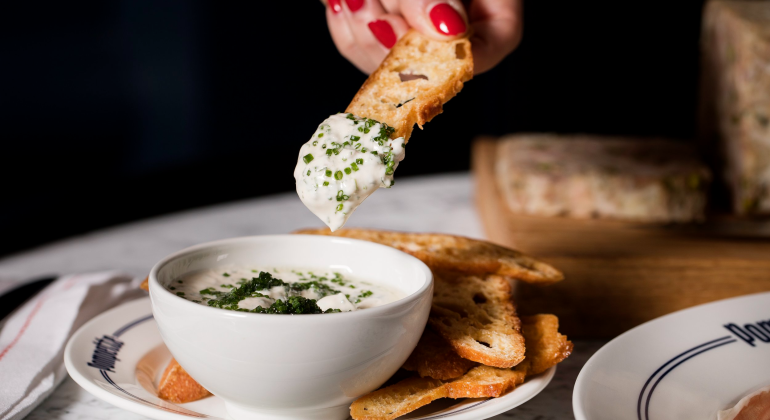 Hand dipping toasted bread into creamy dip with chives on a marble table setting