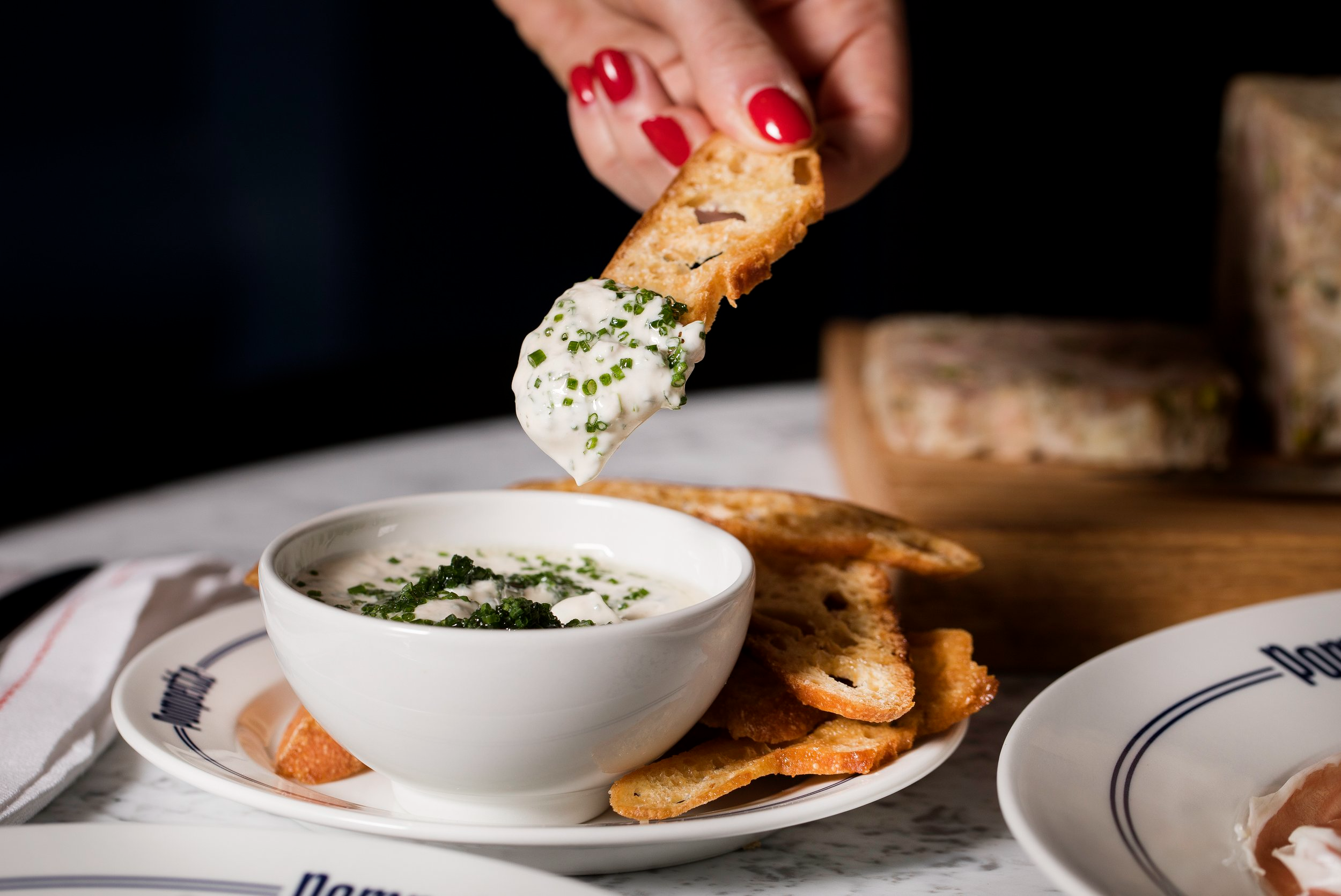 Hand dipping toasted bread into creamy dip with chives on a marble table setting