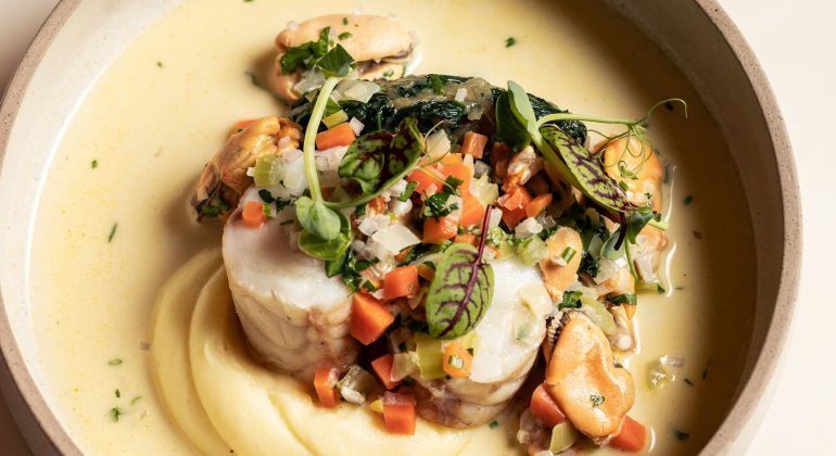 Gourmet dish with seafood, mashed potatoes, and vegetables in creamy sauce in beige bowl on marble table