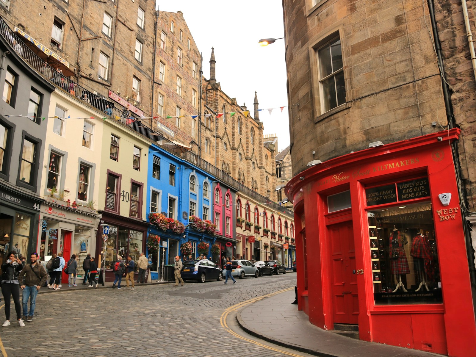 Colourful shops on a historic cobbled street in Edinburgh, featuring a prominent red kilt shop on the corner