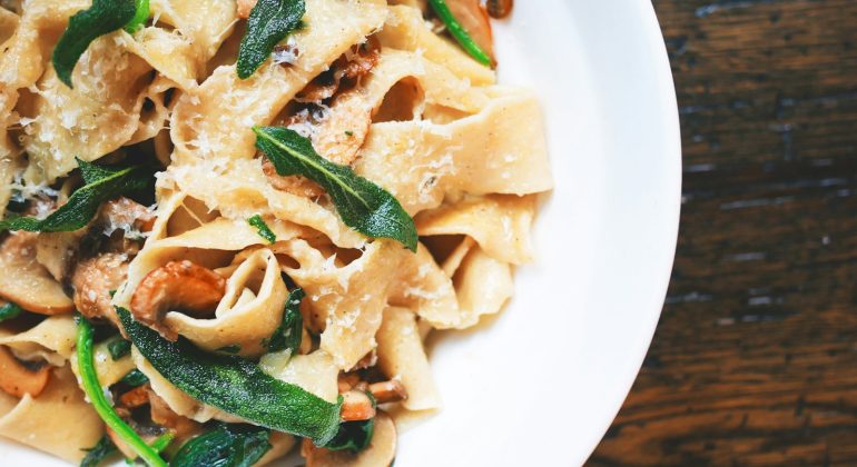 Fresh pappardelle pasta with mushrooms and spinach on a white plate, garnished with herbs and cheese