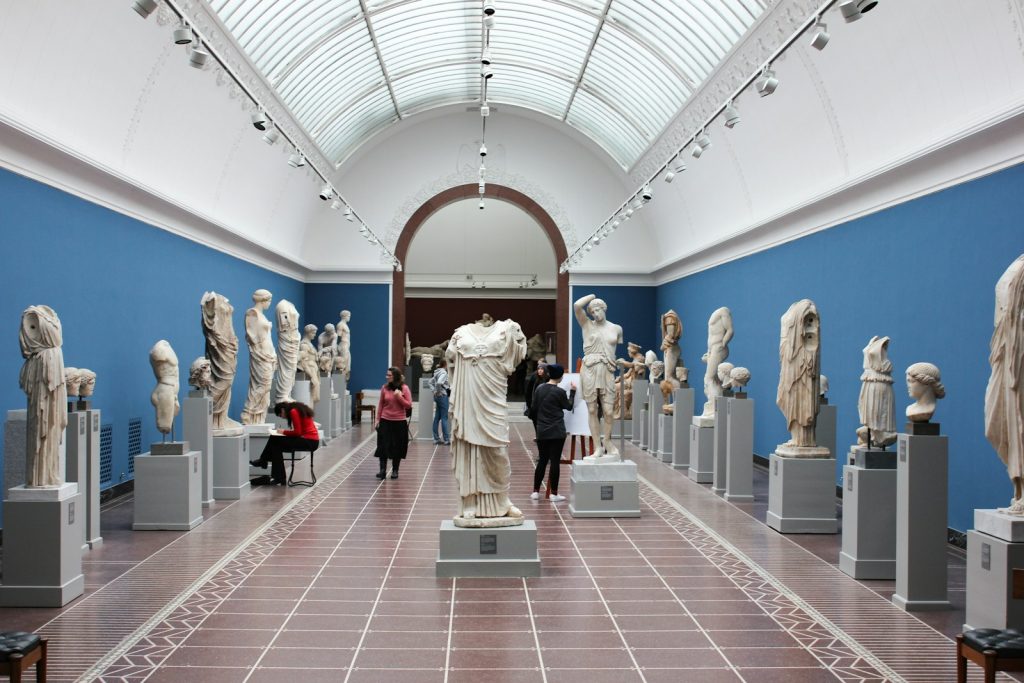 Visitors exploring classical sculptures in a museum gallery with blue walls and high arched ceiling