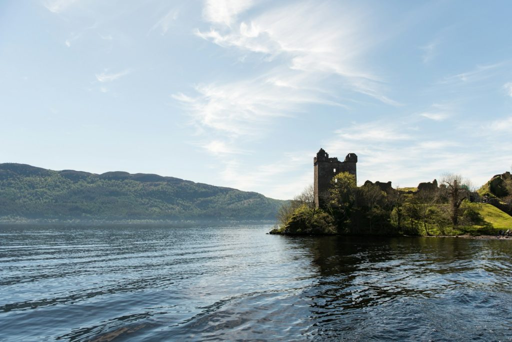 Ancient castle ruins on a hill by a scenic loch with lush green hills in the background under a bright blue sky