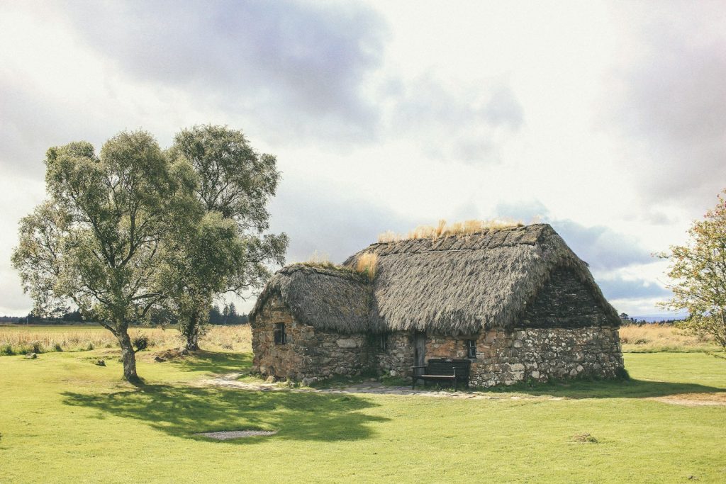 Stone cottage with a thatched roof in a rural landscape surrounded by trees under a cloudy sky in the UK countryside