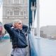 A woman stands on a bridge with Tower Bridge visible in the background, showcasing a scenic urban landscape